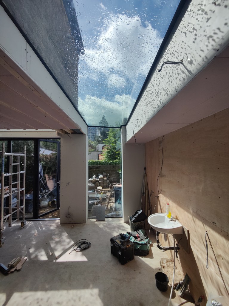 Interior view of a partially constructed room featuring large glass skylights with raindrops, showcasing the exterior garden through glass doors, along with construction materials and tools scattered on the floor.