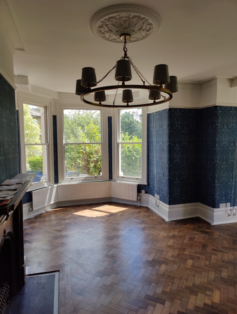 Bright room with a patterned wallpaper, featuring a chandelier and large windows that offer a view of greenery. The floor is made of herringbone parquet.