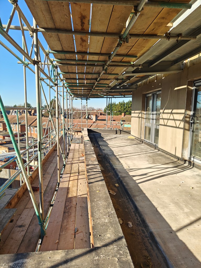 A view of scaffolding on a building site, featuring wooden planks and a concrete surface, with residential rooftops in the background under clear blue skies.