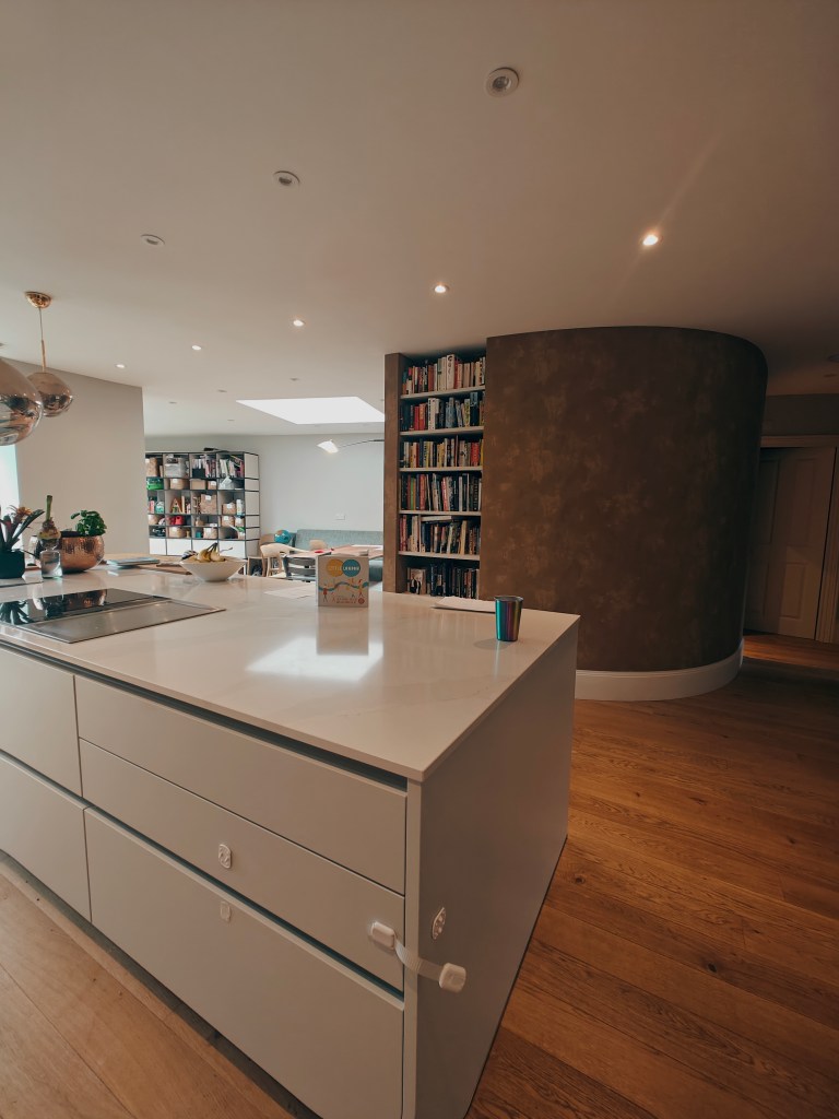 Modern kitchen interior featuring a white island unit with minimalist design, a small colourful cup, and a carton of drink on the countertop. In the background, there is a bookshelf filled with books and a light-filled living area visible through a large window.