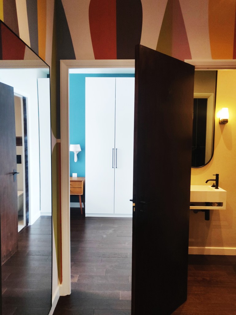 Interior view of a modern room featuring a doorway, a large white wardrobe, a wooden vanity with a round mirror, and a wall adorned with colourful abstract patterns.